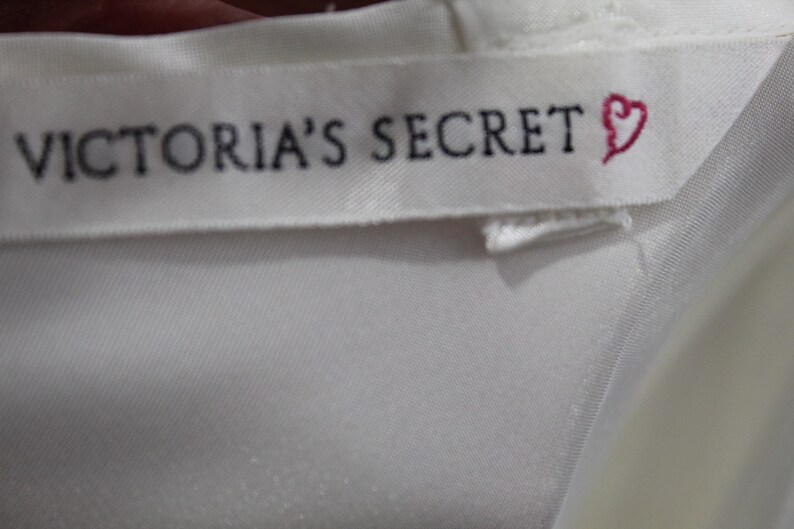 Puede incluir: Primer plano de una etiqueta de tela blanca con el texto "VICTORIA'S SECRET" en negro y un s&iacute;mbolo de coraz&oacute;n rosa. La etiqueta est&aacute; cosida sobre una tela de sat&eacute;n blanca.