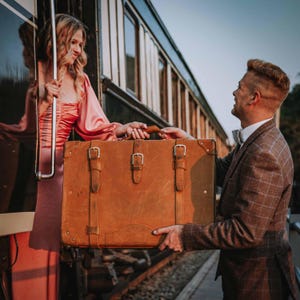 May include: A vintage-style brown leather suitcase with three buckle closures is being held by two people near a train. The suitcase has a rectangular shape and is made of a light brown leather material. The train is dark green.