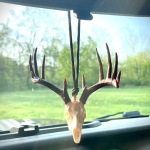 May include: A deer skull car charm with brown antlers and a cream-colored skull. The charm hangs from a camouflage-patterned cord. The background shows a green field and trees, suggesting an outdoor theme.