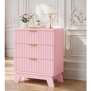 May include: A light pink three-drawer chest featuring a vertical fluted texture on the drawer fronts and slim horizontal gold-toned metal handles. The piece stands on four angled legs and has a flat top surface for storage or display purposes.