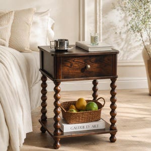May include: A dark brown wooden nightstand with a single drawer and turned legs. A woven basket with fruit sits on the lower shelf. A cup and saucer, a book, and a candle are on top. The nightstand is next to a bed with white bedding.