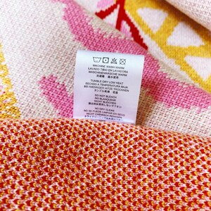 May include: A white tag with black and blue care instructions for a garment. The tag includes instructions for machine washing, tumble drying, bleaching, and dry cleaning. The text on the tag reads: "Machine wash warm, Lavado Tibio en Lavadora, Maschinenw&auml;sche warm, 洗濯機 溫水使用, Tumble dry low heat, Secado a Temperatura Baja, Bei niedriger Hitze trocknen, タンブル乾燥低溫, Do not bleach, No Blanquear, Nicht bleichen, 漂白剤は使用しないで下さい, No dry clean, No se puede limpiar en seco, Nicht chemisch reinigen, ドライクリーニングはできません."