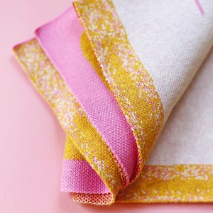 May include: A folded pink and yellow striped fabric kitchen towel with a white background. The towel has a woven pattern and a fringed edge.