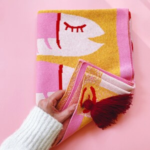 May include: A pink and yellow knitted throw blanket with a white eye design and red accents. The blanket has a red tassel.