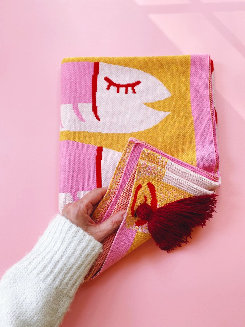 May include: A pink and yellow knitted throw blanket with a white eye design and red accents. The blanket has a red tassel.