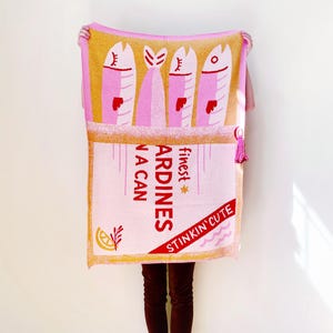 May include: A pink and yellow throw blanket with a design of a can of sardines. The can has the text "finest sardines in a can" and "stinkin' cute" on it.