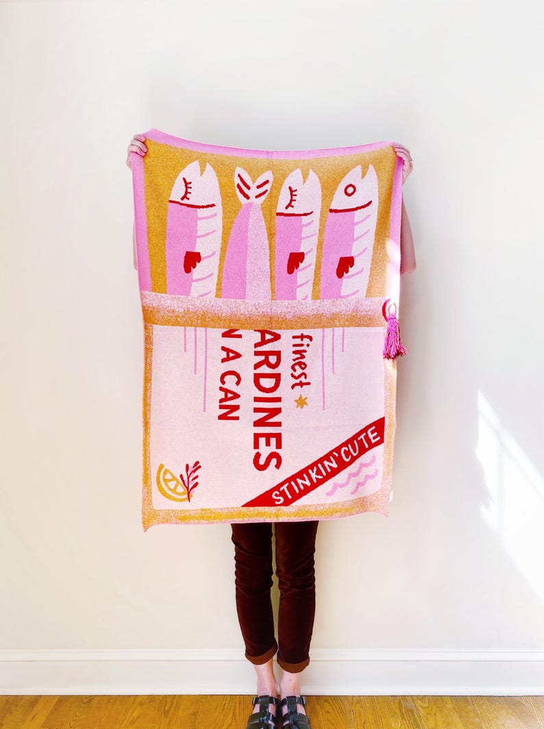 May include: A pink and yellow throw blanket with a design of a can of sardines. The can has the text "finest sardines in a can" and "stinkin' cute" on it.