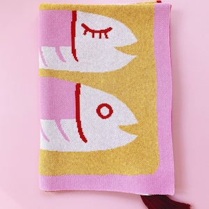 May include: A pink and yellow knitted throw blanket with a red and white fish design. The blanket has a red tassel.