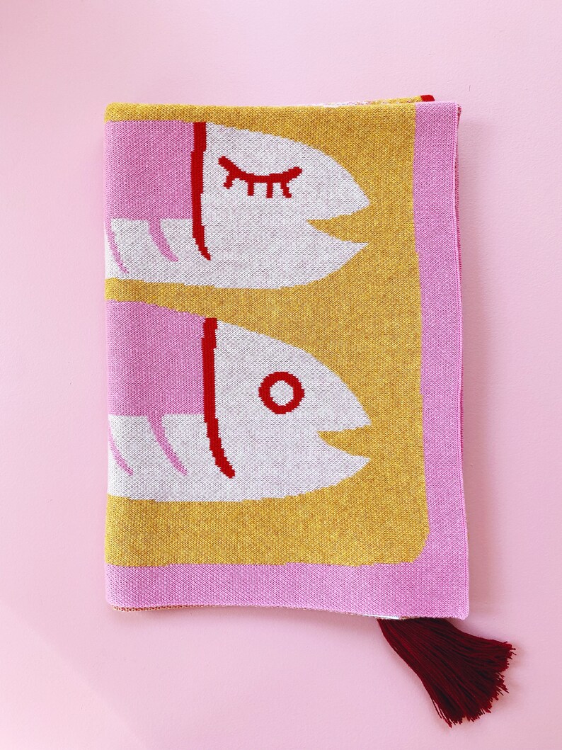 May include: A pink and yellow knitted throw blanket with a red and white fish design. The blanket has a red tassel.