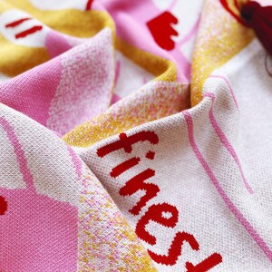 May include: A pink and yellow knitted blanket with a red "finest" text. The blanket has a white background with a subtle pattern.