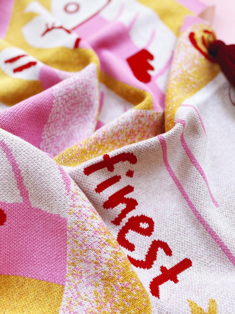 May include: A pink and yellow knitted blanket with a red "finest" text. The blanket has a white background with a subtle pattern.