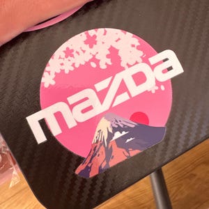 Könnte beinhalten: Ein runder Aufkleber mit einem rosa Sonnenhintergrund, weißen Kirschblütensilhouetten und einer Berggrafik am unteren Rand. Das Wort "mazda" ist in weißen, fetten Kleinbuchstaben in der Mitte des Aufklebers aufgedruckt.