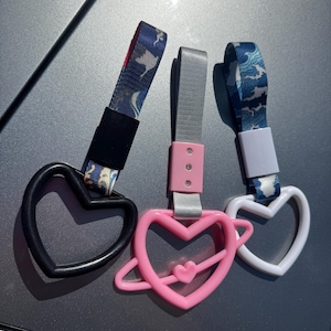 Può includere: Tre accessori per auto a forma di cuore. Uno è nero, uno è rosa con un piccolo cuore al centro e l'altro è bianco. Ognuno ha un cinturino con un design diverso: blu con onde bianche, grigio e blu con onde bianche.
