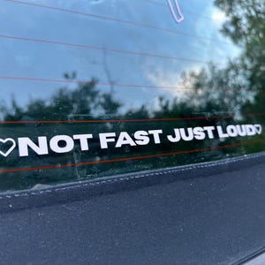 Peut inclure: Autocollant de voiture en vinyle blanc avec le texte "NOT FAST JUST LOUD" en gras, sans empattement. Un symbole de cœur blanc précède le texte. L'autocollant est sur une vitre de voiture, avec un arrière-plan flou d'arbres et de ciel.