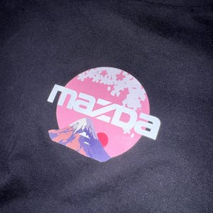 May include: Black t-shirt with a graphic design. The design features the word "MAZDA" in white, set against a pink circle with cherry blossoms. A mountain and red sun are also depicted.