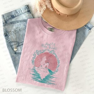 May include: A light pink t-shirt with the text "Part Of Your World" and a graphic of a mermaid in a circle. The shirt is laid on top of blue jeans and a tan hat. A necklace is visible.