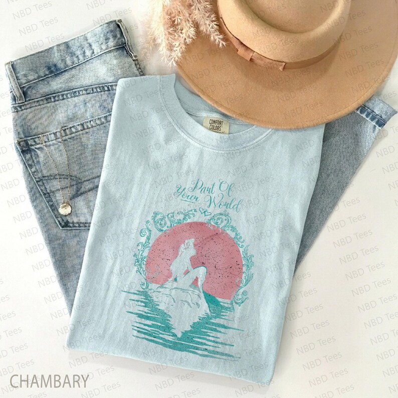 May include: Light blue t-shirt with the text "Part Of Your World" and a graphic of a mermaid silhouette against a pink sun. The shirt is paired with light wash jeans and a tan hat.