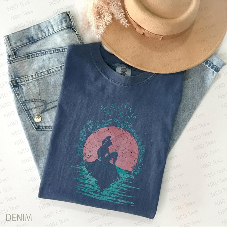 May include: A navy blue t-shirt with the text "Part Of Your World" and a graphic of a mermaid silhouette against a pink and teal background. The shirt is paired with light wash denim jeans and a tan hat.