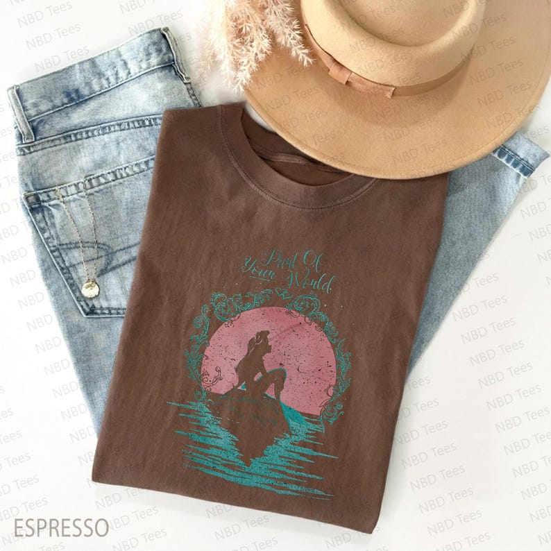May include: A brown t-shirt with the text "Part Of Your World" and a graphic of a mermaid silhouette. The shirt is laid on top of blue jeans, a necklace, and a tan hat.