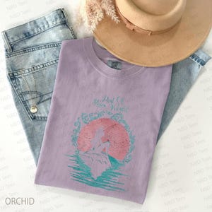 May include: Orchid-colored t-shirt with the text "Part Of Your World" and a graphic of a mermaid in front of a pink sun. The shirt is laid on top of blue jeans and a tan hat. A gold necklace is visible.
