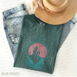May include: A blue spruce t-shirt with a graphic of a mermaid silhouette against a pink sun, with the words "Under the Sea World". The shirt is paired with light wash jeans and a tan hat.