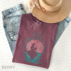 May include: Berry-colored t-shirt with a graphic of a mermaid and the text "Part of Your World." The shirt is paired with light-wash jeans, a gold necklace, and a tan hat. The shirt is a comfortable, casual style.