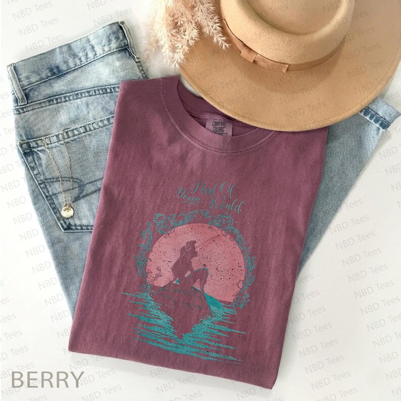 May include: Berry-colored t-shirt with a graphic of a mermaid and the text "Part of Your World." The shirt is paired with light-wash jeans, a gold necklace, and a tan hat. The shirt is a comfortable, casual style.