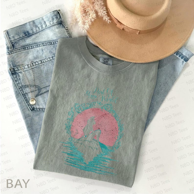 May include: A sage green t-shirt with a graphic of a wolf in front of a pink sun, with the text "Be Wild and Free". The shirt is paired with light wash jeans, a tan hat, and a gold necklace.