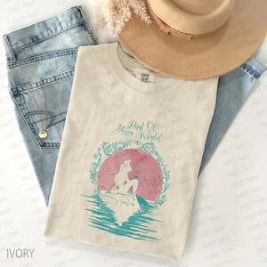 May include: Ivory-colored t-shirt featuring a distressed graphic of a mermaid silhouette in front of a pink sun, with the text "Part Of Your World." The design is framed by a teal decorative border. The shirt is paired with light blue jeans and a tan hat.