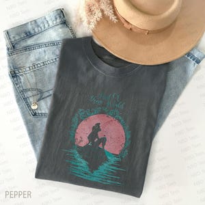 May include: Dark gray t-shirt featuring a teal and pink graphic of a mermaid on a rock with the text "Part Of Your World". The shirt is paired with light blue jeans and a tan hat. A necklace is also visible.