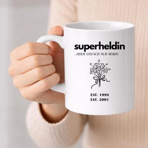 May include: White ceramic mug with the word "superheldin" in black, along with the phrase "...ODER EINFACH NUR MAMA!" and a floral illustration. The mug also features the dates "EST. 1998" and "EST. 2001".