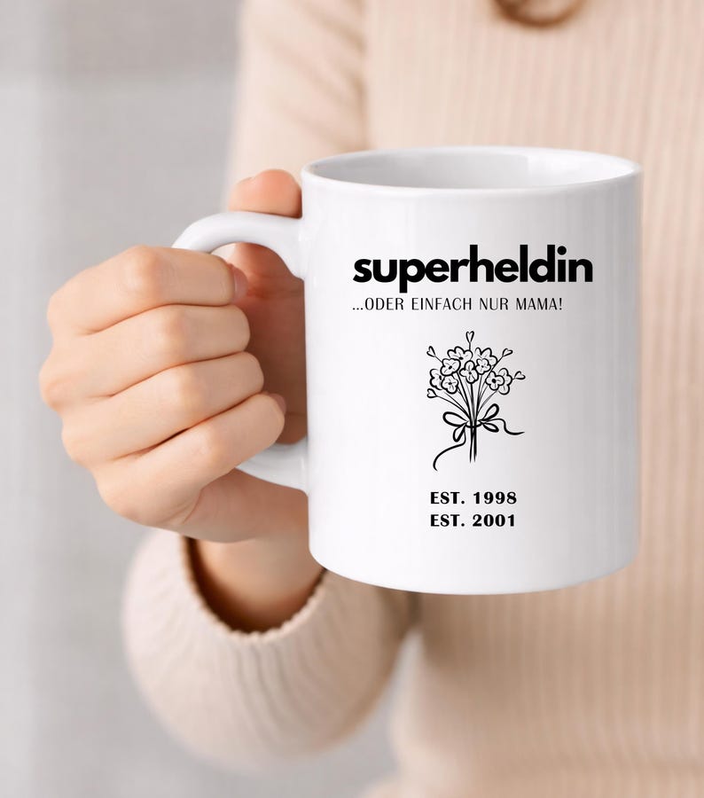 May include: White ceramic mug with the word "superheldin" in black, along with the phrase "...ODER EINFACH NUR MAMA!" and a floral illustration. The mug also features the dates "EST. 1998" and "EST. 2001".