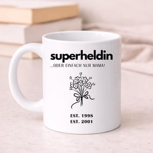 May include: White ceramic mug with the words "superheldin ...ODER EINFACH NUR MAMA!" in black. A black floral design and the dates "EST. 1998" and "EST. 2001" are also featured. The mug has a curved handle.