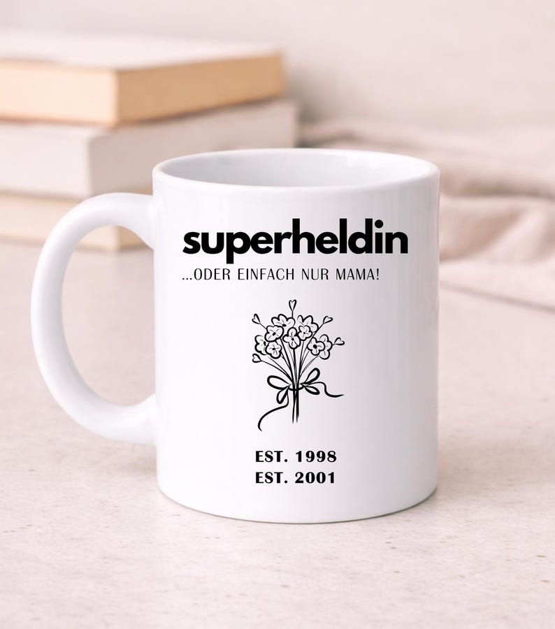 May include: White ceramic mug with the words "superheldin ...ODER EINFACH NUR MAMA!" in black. A black floral design and the dates "EST. 1998" and "EST. 2001" are also featured. The mug has a curved handle.