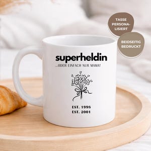 May include: White ceramic mug with the word "superheldin" and the phrase "...ODER EINFACH NUR MAMA!" in black. A floral bouquet graphic and the dates "EST. 1998" and "EST. 2001" are also printed on the mug. The mug is on a wooden tray.