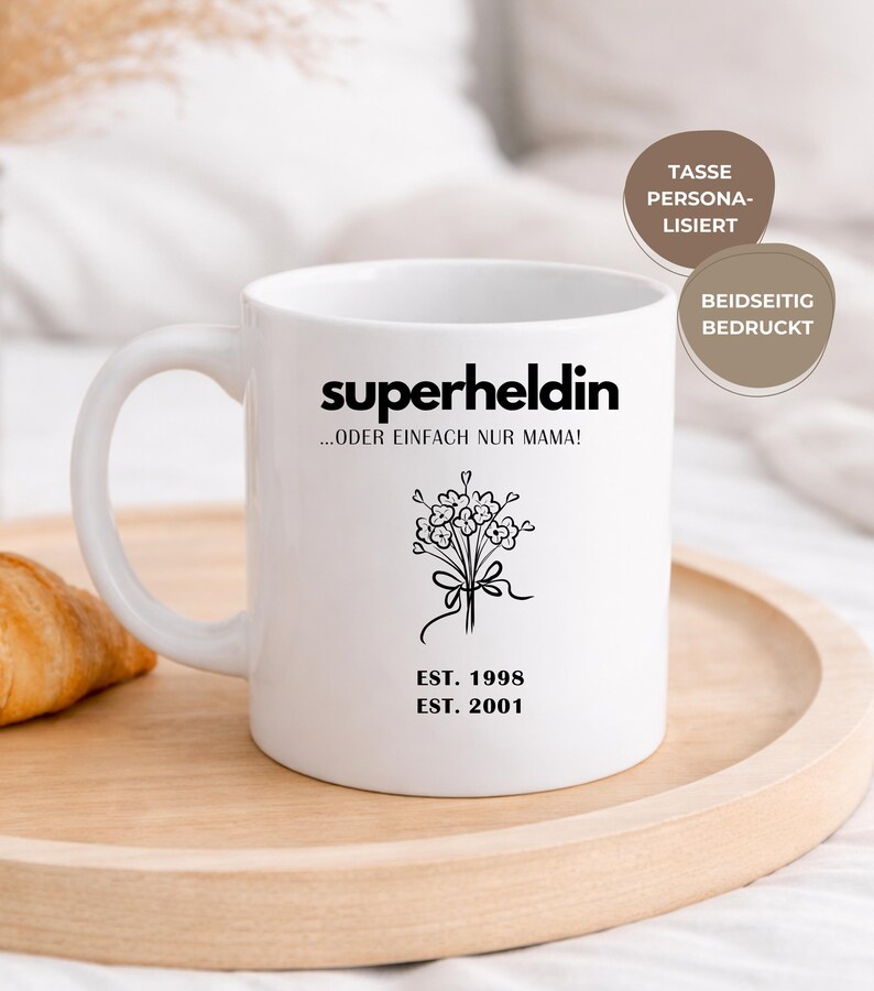 May include: White ceramic mug with the word "superheldin" and the phrase "...ODER EINFACH NUR MAMA!" in black. A floral bouquet graphic and the dates "EST. 1998" and "EST. 2001" are also printed on the mug. The mug is on a wooden tray.