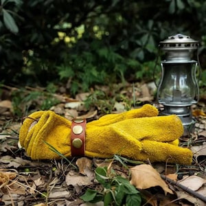 Könnte beinhalten: Senfgelbe Arbeitshandschuhe mit einem braunen Lederarmband, das mit messingfarbenen Nieten befestigt ist. Die Handschuhe liegen auf Blättern, mit einer Vintage-Laterne im Hintergrund.