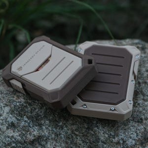 Può includere: Due custodie rettangolari robuste, marroni e beige, appoggiate su una pietra. Le custodie presentano un design strutturato con viti visibili e la scritta "JANSBERRY DESIGN". Sono custodie protettive per dispositivi elettronici.