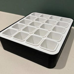 Pode incluir: Um organizador retangular preto e branco com 20 compartimentos quadrados. O organizador tem uma base preta e um topo branco com os compartimentos individuais. Os compartimentos são projetados para guardar itens pequenos.