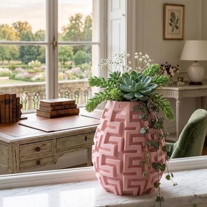 Puede incluir: Un jarrón rosa geométrico con un patrón laberíntico, lleno de vegetación y suculentas. El jarrón está sobre una superficie blanca. El fondo muestra una ventana con vistas a un jardín, un escritorio y una lámpara.