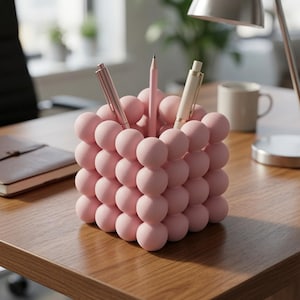 May include: A light pink pen holder constructed from a grid of small, spherical balls. The square-shaped container holds two pens and one pencil, with the matte-textured surface creating a uniform, bubble-like appearance on all visible sides.