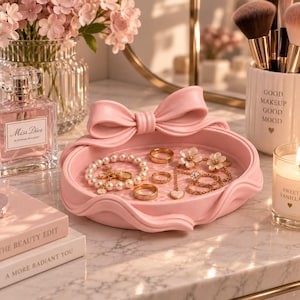 May include: A round, light pink jewellery tray featuring a prominent sculpted bow on the rim. The tray holds a pearl bracelet, three gold-toned rings, and a gold-toned necklace with a small heart pendant and floral-shaped accents.
