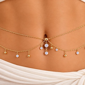 May include: A gold-toned belly chain featuring two delicate layered chains. The lower chain is adorned with small dangling gold spheres and clear round crystals. A central vertical drop element features two additional clear crystals.