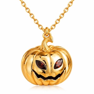 May include: A gold-toned metal necklace featuring a pumpkin-shaped pendant with a carved jack-o'-lantern face. The pumpkin has two dark red, marquise-cut gemstone eyes and a wide, jagged mouth, suspended from a delicate gold-toned link chain.