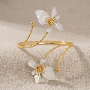 May include: Gold-coloured arm cuff with two white flower accents and gold centres. The cuff has a delicate, floral design with curved, textured arms and small gold spheres at the ends. The flowers have a pearlescent finish.