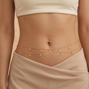 May include: Gold-toned body chain with two strands, each adorned with small, sparkling clear gemstones. The delicate jewelry is draped across the waist, creating a subtle, elegant accessory. The chain is worn over a beige skirt and a white top.