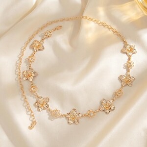 May include: A gold-colored body chain featuring a floral design. The chain is composed of delicate links and flower-shaped pendants. The jewelry is displayed on a white, silky fabric, highlighting its elegant appearance. The chain is designed to be worn around the waist.