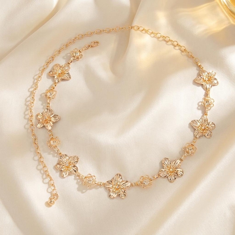 May include: A gold-colored body chain featuring a floral design. The chain is composed of delicate links and flower-shaped pendants. The jewelry is displayed on a white, silky fabric, highlighting its elegant appearance. The chain is designed to be worn around the waist.