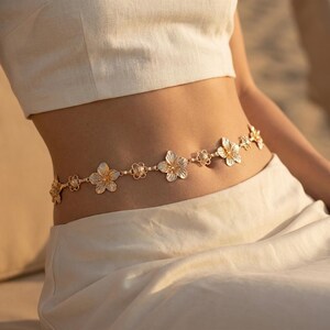 May include: A gold-toned waist chain featuring a floral design. The chain is composed of delicate flower-shaped links and smaller, circular links. The flowers have a detailed, textured appearance, and the chain is worn around the waist.