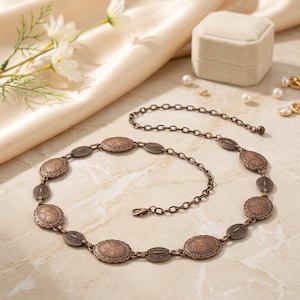 May include: A bronze-toned metal belt with oval and circular decorative elements. The belt features an intricate pattern on each oval and circular piece, connected by a chain. The belt is displayed on a light-colored surface.